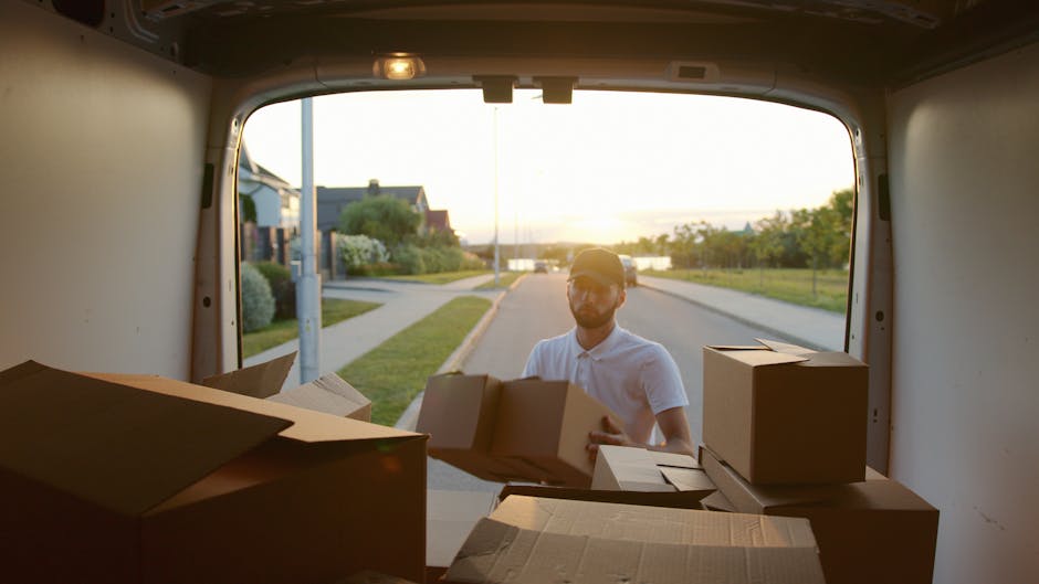 A man wearing a white t-shirt and glasses stands on a residential street, carefully stacking cardboard boxes outside a white van with its rear doors open. The boxes are of varying sizes, some taped an