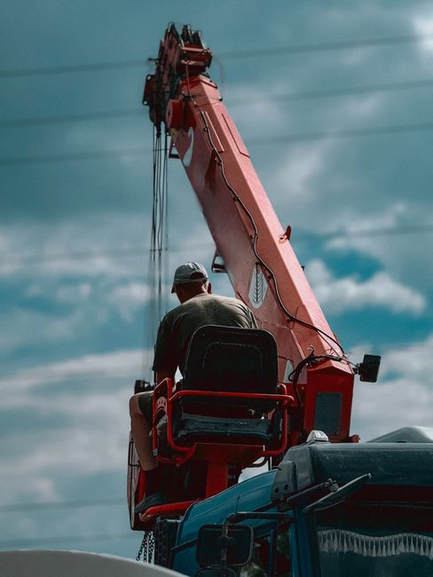 A man wearing a grey cap and dark clothing is seated on the control platform of a large red crane with a telescopic arm extended upwards, set against a cloudy sky. The crane is mounted on a vehicle, with visible parts of the vehicle's blue body in the foreground. The scene is captured outdoors, with the crane appearing to be in use for lifting or moving large objects, possibly related to home relocation or furniture transport. Man with Van Ilford specializes in removals, including tasks that require crane access for bulky items or difficult-to-reach areas, as part of their house removal services on Cranbrook Road.