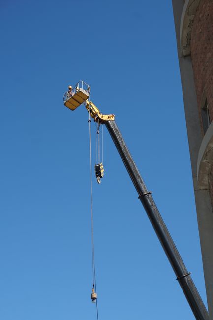 A crane with a yellow telescopic arm is extended high into a clear blue sky, lifting a rectangular yellow work platform with construction workers wearing safety helmets and harnesses. The crane's arm is attached to a hook and pulley system that supports the platform, which is positioned adjacent to a building's exterior wall. The building features a curved brick facade and has multiple arched openings, indicating an architectural style. The scene appears to be part of a home relocation or building maintenance operation, with the crane used to facilitate the lifting of bulky items or materials to higher floors. The image captures the active process of furniture transport or bulk item removals, essential components of professional house removals, with visible safety measures and equipment such as the crane, pulleys, and harnesses. This setup aligns with industry standards for safe, efficient packing and moving logistics, as seen in the services offered by Man with Van Ilford on Cranbrook Road.