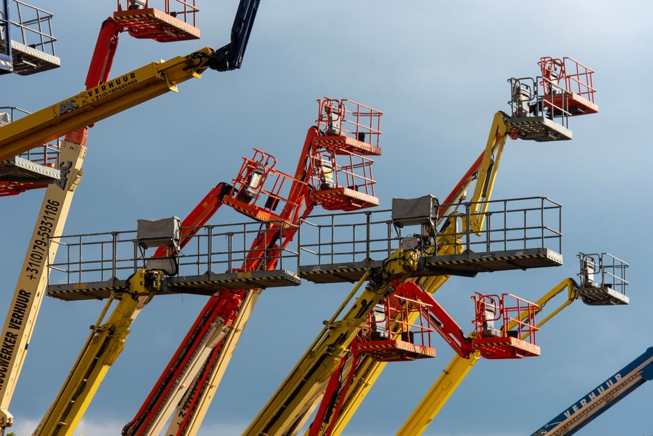 A man wearing a grey cap and dark clothing is seated on the control platform of a large red crane with a telescopic arm extended upwards, set against a cloudy sky. The crane is mounted on a vehicle, with visible parts of the vehicle's blue body in the foreground. The scene is captured outdoors, with the crane appearing to be in use for lifting or moving large objects, possibly related to home relocation or furniture transport. Man with Van Ilford specializes in removals, including tasks that require crane access for bulky items or difficult-to-reach areas, as part of their house removal services on Cranbrook Road.
