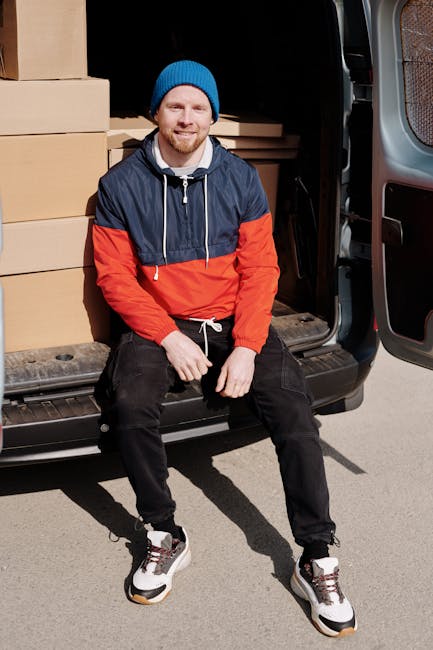 A man with a beard, wearing a blue knitted hat and a color-blocked jacket in navy blue and bright orange, is sitting on the edge of the open rear door of a black moving van. Inside the van, there are multiple cardboard boxes of various sizes neatly stacked, some wrapped in plastic film. The boxes are positioned on the van's floor, with additional packing materials visible nearby. The man is smiling at the camera with one hand resting on his knee and the other on a box. The surrounding environment appears to be an outdoor area with pavement, and the scene is well-lit with natural daylight. This image depicts the process of furniture transport and packing during home or business relocation, with Man with Van Ilford providing a professional removals service in accordance with their house removals and moving logistics offerings.