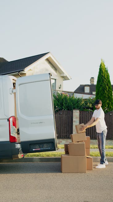 A man stands outside a residential property next to a white MAN WITH VAN ILFORD vehicle during a home relocation process. He is wearing a white T-shirt and jeans, and is in the act of unpacking or packing cardboard boxes, which are placed on the pavement beside him. The open rear door of the van reveals additional cardboard boxes and packaging materials inside, suggesting the ongoing loading or unloading process involved in furniture transport or house removals. The scene occurs in a suburban neighbourhood with neatly maintained gardens, a wooden fence, and a house with a dark roof visible in the background. Bright natural daylight illuminates the scene, emphasizing the typical steps of packing, loading, and managing moving logistics in a professional removal service. Man with Van Ilford specializes in local removals, supporting efficient and careful packing and transport for house or office moves.