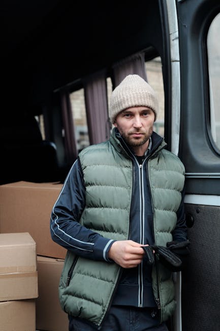 A man with a beard, wearing a white polo shirt, black cap, and jeans, standing inside the open back of a commercial moving van filled with stacked cardboard boxes of various sizes, some wrapped in pla
