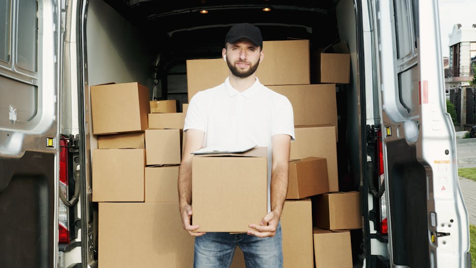 A man with a beard, wearing a black cap, white polo shirt, and jeans, stands inside the open rear of a white moving van filled with stacked cardboard boxes in various sizes, some wrapped in plastic or packing paper. He holds a medium-sized box with both hands, positioned at waist height. The van is parked on a driveway near a residential area, with a house and greenery visible in the background. The scene depicts the loading process involved in house removals and furniture transport, with the interior of the van showing clean and organized space for home relocation purposes. Man with Van Ilford appears to be preparing or assisting with packing and moving activities, emphasizing professional packing, logistics, and transport of household items.