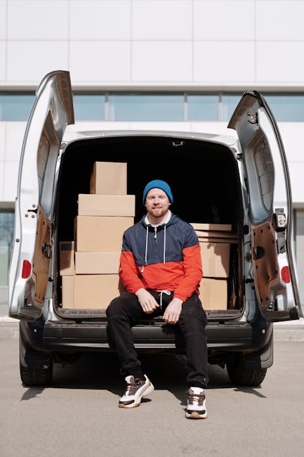 A man with a beard, wearing a black cap, white polo shirt, and jeans, stands inside the open rear of a white moving van filled with stacked cardboard boxes in various sizes, some wrapped in plastic or packing paper. He holds a medium-sized box with both hands, positioned at waist height. The van is parked on a driveway near a residential area, with a house and greenery visible in the background. The scene depicts the loading process involved in house removals and furniture transport, with the interior of the van showing clean and organized space for home relocation purposes. Man with Van Ilford appears to be preparing or assisting with packing and moving activities, emphasizing professional packing, logistics, and transport of household items.