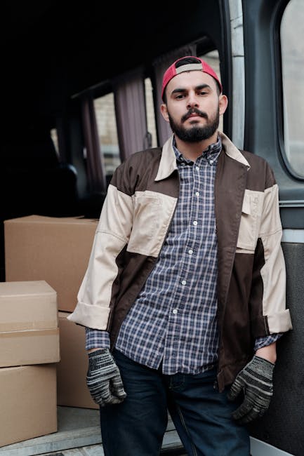 A young man with a beard, wearing a red cap backwards, a brown and beige jacket over a blue checked shirt, and grey patterned gloves, is sitting on the edge of an open van's cargo area during a house removal. Behind him inside the vehicle, there are several large cardboard boxes, some stacked vertically and others placed side by side, ready for transportation as part of a home relocation process. The van has a dark interior with a partially visible driver's seat and headrest. The man appears to be taking a break during packing and loading activities, with a neutral facial expression. The environment suggests an organized loading process, with the boxes secured in the rear of the vehicle. This scene illustrates furniture transport and packing-for-move preparations undertaken by a professional removals service such as Man with Van Ilford, as part of their house removals and moving logistics services, connected to Redbridge Council removals permits in Ilford.
