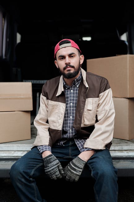 A young man with a beard, wearing a red baseball cap, a checkered shirt, and a two-tone beige and brown jacket, is sitting on the edge of an open van trunk. He has grey and black striped gloves on both hands, which rest on his knees. Behind him inside the van, there are several cardboard boxes of various sizes stacked on the vehicle's floor. The boxes are sealed with packing tape and appear ready for transportation as part of a house removal or home relocation process. The van's interior is dark, with a visible headrest of a vehicle seat behind the young man. The scene reflects the loading or packing stage involved in furniture transport and moving logistics, with the focus on careful handling of belongings typical for same-day or emergency moves facilitated by Man with Van Ilford.