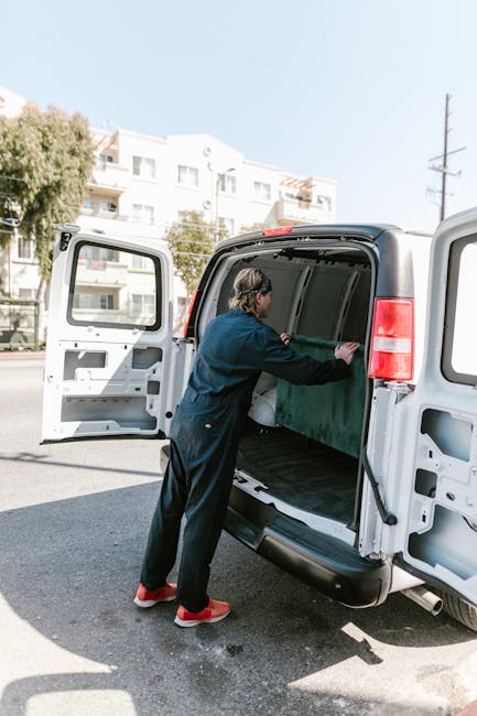A person wearing a dark jumpsuit and red sneakers is loading or unloading a large, green wooden crate into the rear cargo space of a white panel van parked on a street. The van's side and rear doors are open, revealing the empty interior lined with protective padding. In the background, there are multi-story residential buildings with balconies, trees, and utility poles under a clear blue sky. The scene depicts a home relocation or furniture transport process, with the individual carefully handling items during a same-day moving service. Natural lighting highlights the details of the scene, and the company Man with Van Ilford is known for providing removals and moving logistics.