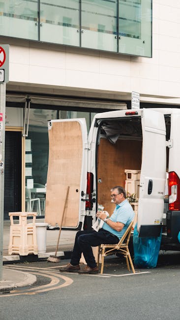 A man with glasses, wearing a light blue shirt and dark trousers, is seated on a wooden chair beside a white moving van parked on a narrow street. The van's rear doors are open, revealing an interior