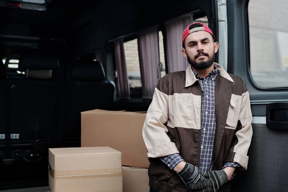 A man with a beard, wearing a red cap backwards, a checkered shirt, and a beige and brown jacket, stands inside the open rear of a van used for home relocation services. The van's interior features partially visible black seats and a window with a light-colored curtain. Next to the man are several cardboard boxes in various sizes, some sealed with packing tape, prepared for packing and moving. The scene is outdoors, and the van is parked on a paved surface, with a glimpse of a brick building visible through the van's side window. The man appears to be part of a professional removals team associated with Man with Van Ilford, providing furniture transport and packing services during a house move, aligned with the context of Valentines Park home removals in Ilford.